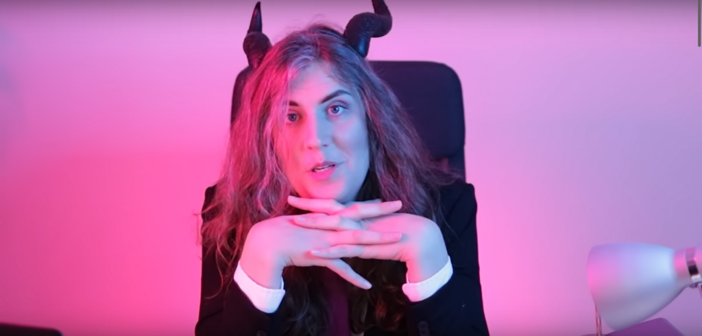 YouTuber*in Verity Ritchie von verilybitchie sitzt mit Teufelshörnern auf dem Kopf in einem Chefsessel und hat die Hände unterm Kinn gefaltet. Sie ist in pinkes und violettes Licht getaucht, vermutlich eine Anspielung auf die bisexuelle Flagge.